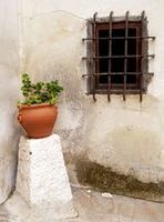 Fachada de una casa. Es una pared blanca en la que puede verse una pequeña ventana enrejada y una gran mancha de humedad, en primer plano vemos un pequeño pedestal con una maceta de cerámica que tiene una planta.