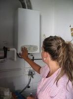 Esther (22 años rubia de pelo largo recogido en una coleta y uniforme de TAPD de rayas rosas y blancas) está accionando los botones de un calentador de agua a gas. Se ve de perfil.