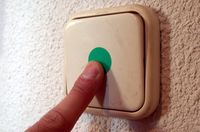 Primer plano de un interruptor que está fijo en una pared blanca. Es de color blanco en el centro tiene un punto de color verde y un dedo lo está accionando.