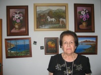 Primer plano de María. Una mujer de 79 años. Está mirando fijamente a la cámara. Le gusta mucho arreglarse y ponerse guapa, tiene el cabello corto, teñido de negro y aparece incluso con los labios pintados. Viste camiseta negra. En la pared de detrás están colgados muchos cuadros que ella misma pinta.