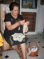 Primer plano de María. Una mujer de 79 años. Está mirando fijamente a la cámara. Le gusta mucho arreglarse y ponerse guapa, tiene el cabello corto, teñido de negro y aparece incluso con los labios pintados. Viste camiseta negra y falda negras. Está sentada en su butaca favorita haciendo croché. En la pared de detrás están colgados muchos cuadros que ella misma pinta.