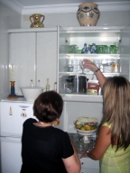 En la cocina de María. María de perfil, mujer de 72 años vestida de negro. Señala a Esther que coja una copa de cristal de uno de los muebles altos de la cocina que permanece abierto. Esther (chica 22 años, pelo rubio y largo y vestido verde) de espaldas a la cámara estira el brazo para coger la copa.