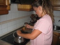 Esther. Una chica de 22 años. Mira a la cámara con una gran sonrisa. Es rubia con el pelo largo, lo tiene recogido hacia atrás y rizado con espuma. Tiene dos perlas grandes blancas como pendientes y viste uniforme a rayas rosas y blancas. Está de pie en la cocina, junto a la ornilla vitrocerámica destapando una olla.