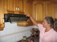 Esther 22 años, rubia, pelo largo recogido en una coleta y uniforme de TAPD de rayas rosas. De pie en la cocina, está accionando los botones de la campana extractora.