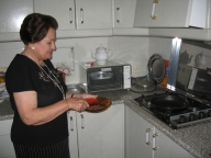 En la cocina de María. María de perfil, mujer de 72 años vestida de negro. Sostiene entre sus manos un plato y un cubierto frente a la hornilla.