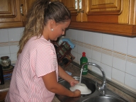 Esther 22 años, rubia, pelo largo recogido en una coleta y uniforme de TAPD de rayas rosas. Está de pie en la cocina, junto al fregadero, lavando la vajilla.