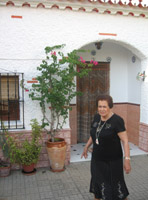 María. Una mujer de 79 años. Le gusta mucho arreglarse y ponerse guapa, tiene el cabello corto, teñido de negro y aparece incluso con los labios pintados. Viste camiseta negra. Camina por delante de la fachada de su casa.