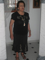 María  mujer de 79 años con el cabello corto, teñido de negro. Y un vestido también negro. Permanece de pie en el pasillo de su casa.