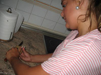 Esther. Una chica de 22 años. Es rubia con el pelo  recogido hacia atrás. Tiene dos perlas grandes blancas como pendientes y viste un uniforme a rayas rosas y blancas. Pela una patata sobre la encimera de la cocina.