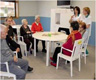 Grupo de personas mayores sentadas alrededor de una mesa, escuchando a dos personas jóvenes trabajadoras del Centro Residencial que están de pie. Se trata de una sesión informativa o formativa.