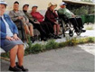 Grupo de personas mayores sentadas en el exterior de un centro residencial. Todos llevan sombreros y gorras para protegerse del sol en lo que puede ser un día de primavera.