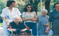 n el centro de la foto aparece una mujer anciana en silla de ruedas. Le guía la silla de ruedas una persona con traje de trabajo blanco de un centro residencial. Al lado le acompaña en el camino 3 mujeres más de distintas edades, familiares de la persona usuaria de la silla de ruedas.