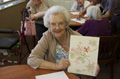 Aparece una señora anciana sonriente, mostrando  orgullosa un cuadro que ha debido realizar ella.