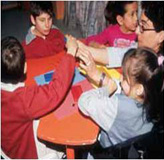 En la imagen aparecen un grupo de niños y niñas sentados alrededor de una mesa. Uno de los niños levanta la mano y la profesora sentada frente a él le sostiene el brazo intentando que le baje.