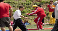 En la imagen aparece un  grupo de personas realizando deporte en el exterior en una pista deportiva. En el centro de la imagen aparece una joven con discapacidad intelectual vestida con chandal rojo y gorra amarilla intentando saltar a la pata coja.