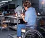 En la foto aparece una mujer de espaldas, usuaria en silla de ruedas, trabajando en un taller.