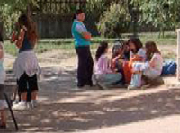 En la imagen aparecen un grupo de chicas jóvenes sentadas en  el suelo de un parque. Una de estas chicas está frente a ellas con los brazos cruzados y de pie.