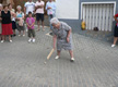 En la imagen aparece una señora mayor jugando en la calle a un juego tradicional, mientras el resto de las personas le observan atentamente.