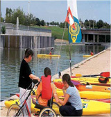 En la imagen aparece un pantano donde se realizan actividades de piraguas. Una persona usuaria de silla de ruedas aparece en la imagen ayudada por otras dos personas.