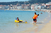 En la imagen se ve a un joven con discapacidad motora utilizando una silla anfibia en la playa y ayudado por dos socorritas.