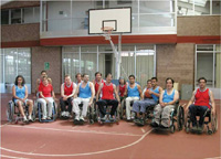 En la imagen aparece un grupo de personas usuarias de sillas de ruedas vestidos con ropa deportiva de baloncesto. Detrás de ellos hay una canasta de baloncesto.