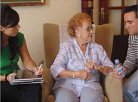 En la imagen aparece una señora mayor en un sillón en el salón de su domicilio y dos profesionales que le están haciendo preguntas, un chico y una chica, esta última recoge la información apuntando en un cuaderno.