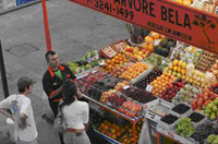 Un puesto en la calle de frutas y verduras con el vendedor atendiendo a dos personas.