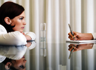 Imagen de una mujer apoyada sobre sus manos en una mesa de cristal, donde se aprecia su reflejo, frente a otro hombre, del que sólo se ve su mano, realizando una serie de anotaciones sobre un papel.