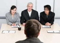 Entrevista de trabajo. Representación de una entrevista de trabajo de un aspirante y tres entrevistadores.La foto está tomada desde la espalda del entrevistado, del que se ve sólo la parte de atrás de su cabeza, y frente a él, en una mesa, con documentación delante de ellos, hay un hombre con el pelo canoso vestido de traje y dos mujeres a sus lados, también de traje, observándole con las manos entrelazada y apoyadas sobre la mesa.