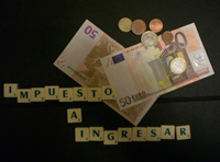 La imagen muestra una fotografía de dinero (billetes y monedas), y en unas fichas con letras verdes sobre fondo blanco dice, “impuesto a ingresar”, todo sobre fondo en negro.
