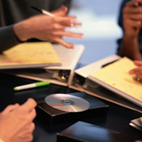 En la imagen se ven: un DVD, unos documentos y las manos de personas que parecen reunidas alrededor de una mesa, y que depositan lo anterior sobre ella. 