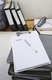 Aparece en primer plano un documento en blanco con un bolígrafo sobre una cartera de documentos, en segundo plano varias carpetas clasificadoras de documentos.