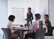 Aparecen cuatro personas dos hombres y dos mujeres, tres de ellos aparecen sentados alrededor de una mesa redonda y una mujer de pie delante de una pizarra de papel exponiendo o explicando.