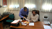 Fotografía de dos hombres en un despacho, Juan sentado junto a una mesa y Pedro al lado hablándole.