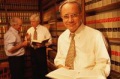 Tres hombres en un despacho de abogados. Uno de ellos lleva camisa blanca y  corbata, y está mirando un libro.