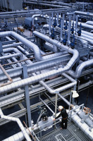 Tipos distribución. Vista superior de una industria, con una gran cantidad de tuberías de diferentes diámetros circulando en todas dimensiones. También aparece un operario con un casco blanco y tamaño en comparación con las tuberías es muy pequeño.