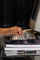 Unas manos introducen datos en un ordenador a través de un teclado de un ordenador, el monitor se observa de lado. El ordenador está encima de una mesa con una documentación y unos bolígrafos.