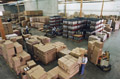 Un almacén visto desde arriba con gran cantidad de productos almacenados en cajas de color marrón formando. Montones. Hay varios operarioa trabajando y uno de ellos conduce una carretilla eléctrica cargada de cajas.