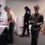 Tres personas en la sala de fotocopiadoras de una empresa, dos de las personas están haciendo fotocopias, la otra  con documentación en la mano.