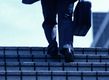 Aparecen las piernas de un hombre que lleva un maletín vestido con pantalón largo gris subiendo unas escaleras y visto desde atrás y de abajo hacia arriba.