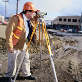 Tipos de carreteras. Aparece un hombre vestido con ropa de trabajo y casco en una carretera haciendo una medición topográfica.