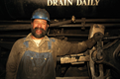 Aparece un hombre con barba, vestido con ropa de trabajo, lleva un mono azul y un casco. Está en un taller  y una de las manos la tiene apoyada en una máquina.