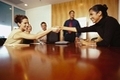 Aparecen dos mujeres  estrechando la mano sentadas alrededor de una mesa de trabajo con tres personas más.