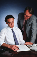 Normas. Dos hombres sentados con camisa y corbata consultan un documento sobre una mesa de trabajo. Uno de los hombres está sentado y el otro está de pie y tiene chaqueta y gafas. Ambos hombres consultan con actitud concentrada el documento.