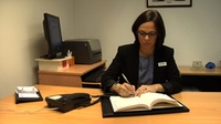 Responsable de comercialización. Mujer con pelo corto y gafas, sentada en una mesa de oficina escribiendo algo.