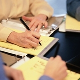 Dos hombres a los que solo se le ve las manos están redactando una documentación en unas hojas de color amarillo, encima de una mesa.