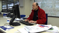 Hombre de unos cuarenta y cinco años sentado delante de un ordenador trabajando. Dispone de una mesa amplia que tiene llena de documentación. Está vestido de forma informal.