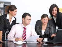 Fases del equipo 3. Tres mujeres y un hombre, todos elegantemente vestidos, miran algo en un portátil, mientras sostienen algunos documentos en la mano. Sobre la mesa hay un par de tazas de café.