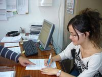 Blanca firmando su contrato de trabajo. Imagen con un plano medio de Blanca, sentada en una mesa, firmando papeles, mientras se ve la mano de alguien que le va indicando con el dedo algún detalle del documento. ”