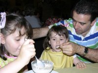 Hombre cuidando a sus hijas. Imagen que muestra a un hombre y a sus dos hijas. Una de las hijas está comiendo por sí sola, pero la otra es demasiado pequeña. La imagen muestra al padre dándole de comer a la niña.